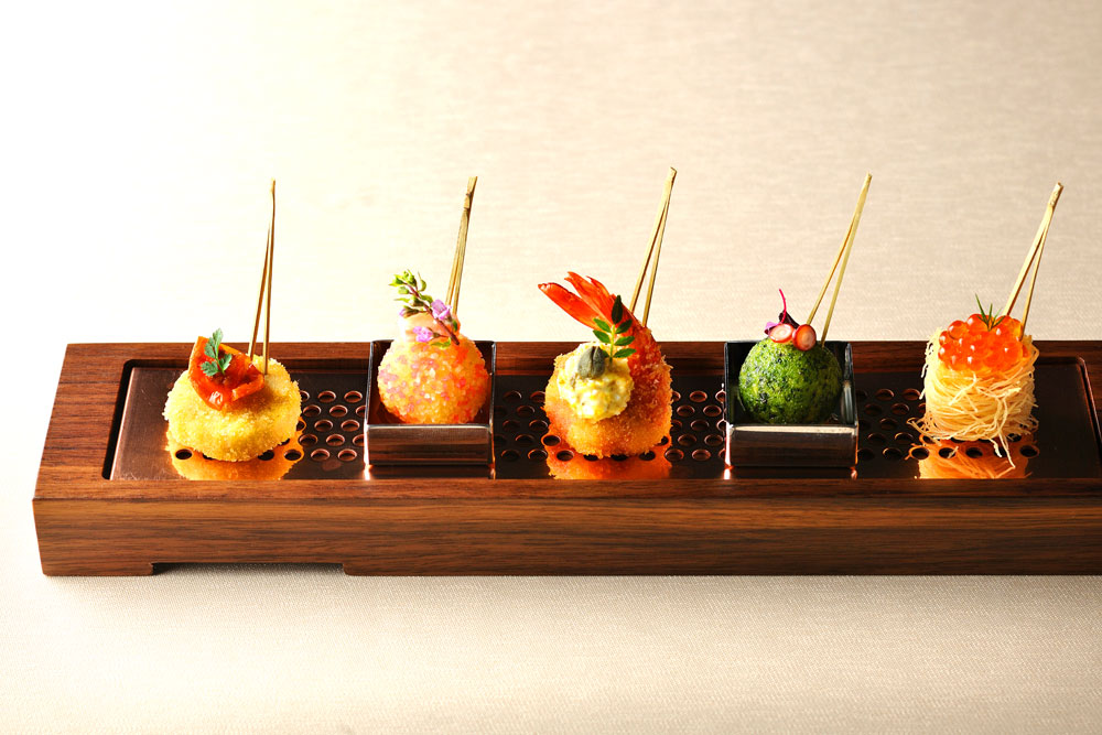 「Naniwa KUSHI Cuisine（なにわ串キュイジーヌ）」の一品。軽井沢の「ブレストンコート ユカワタン」時代から、浜田氏のシグネチャースタイルである“小さなコース”は串カツのプレゼンテーションにも。貝のムースやアメリケーヌソース、ロメスコソースなど、串カツというジャンルにフレンチの要素を加えることで“大阪＆洋”のフュージョンテイストに仕上げている